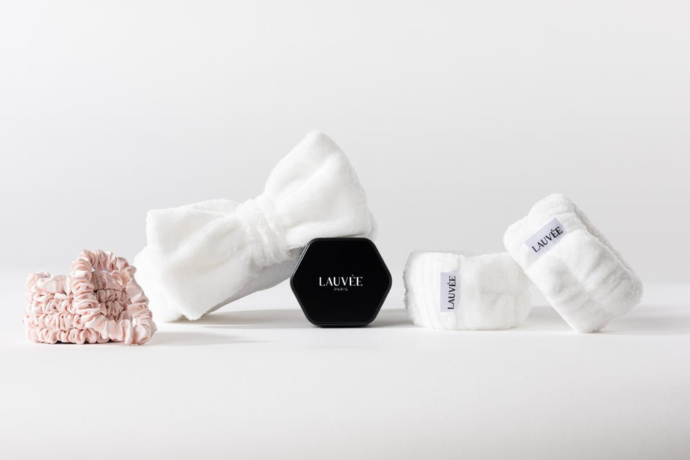 Flat lay sur fond blanc de plusieurs accessoires de soin : un bandeau chenille blanc, deux bracelets essorage blancs marqués Lauvée, un chouchou en soie rose et un boîtier hexagonal noir portant le logo Lauvée Paris.