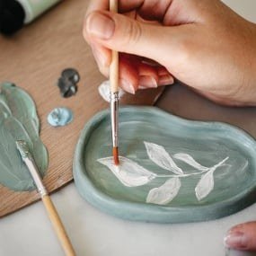 Main peignant au pinceau un motif de feuilles blanches sur une coupelle verte