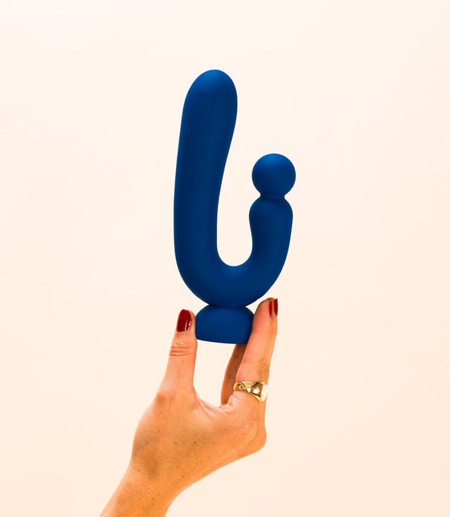 Une main tient un stimulateur intime bleu marine en forme de U de la marque Puissante sur fond beige clair.