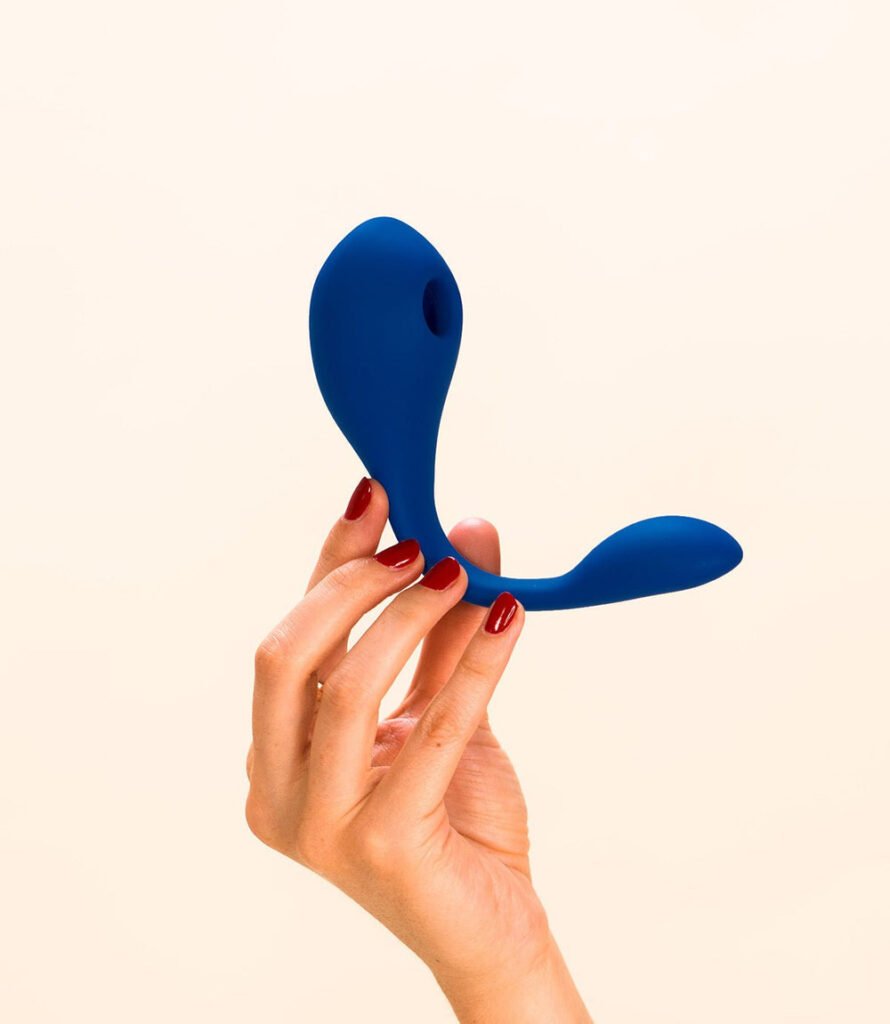 Une main aux ongles rouges tient un stimulateur intime bleu marine de forme organique de la marque Puissante sur fond beige clair.