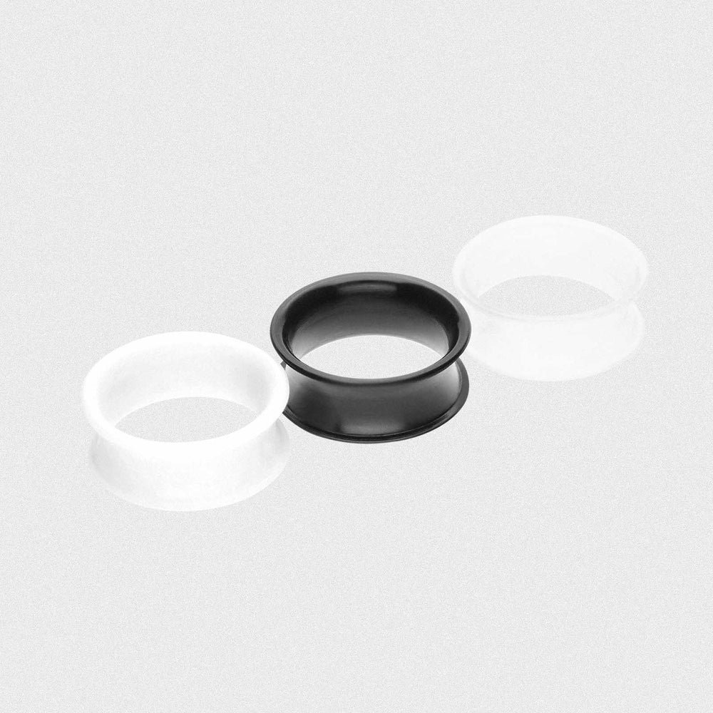 Trois tunnels double flare en silicone — blanc, noir et transparent — posés sur un fond gris neutre.