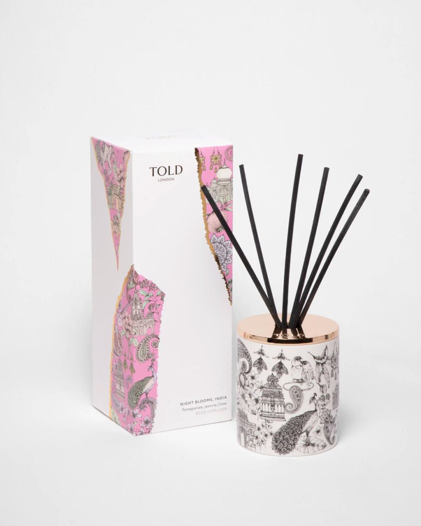 Diffuseur à tiges Night Blooms India dans son pot en porcelaine illustré avec couvercle rosé et tiges noires, posé devant son coffret d'emballage Told London rose et blanc.